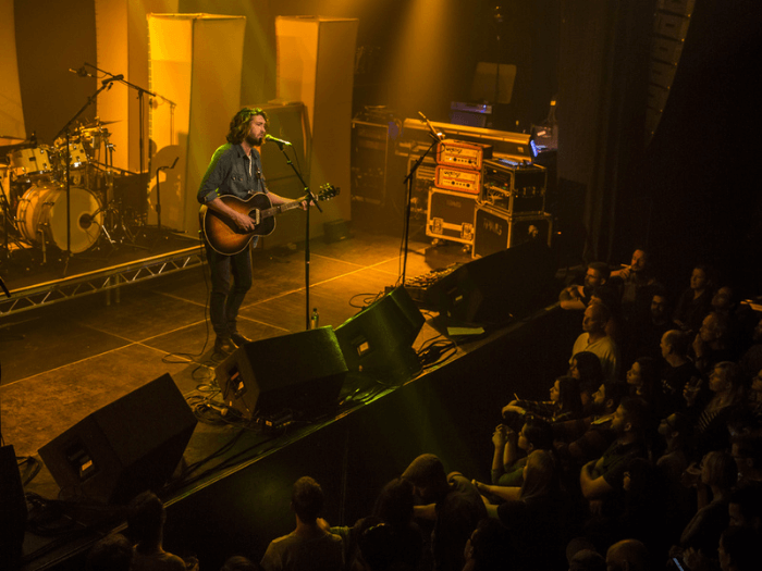 A musician with an acoustic guitar singing into a microphone on stage at a music venue, as audience members watch.